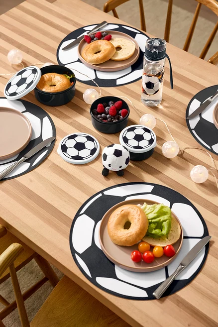 Na drewnianym stole leżą cztery podkładki, bidon, trzy pojemniki na lunch, pluszowa piłka nożna z dźwiękiem i łańcuch świetlny LED »Piłka nożna«.