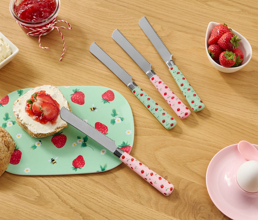 Na drewnianym stole znajdują się bułka, talerz we wzór truskawek, serek śmietankowy, dżem, truskawki i cztery noże we wzór truskawek.