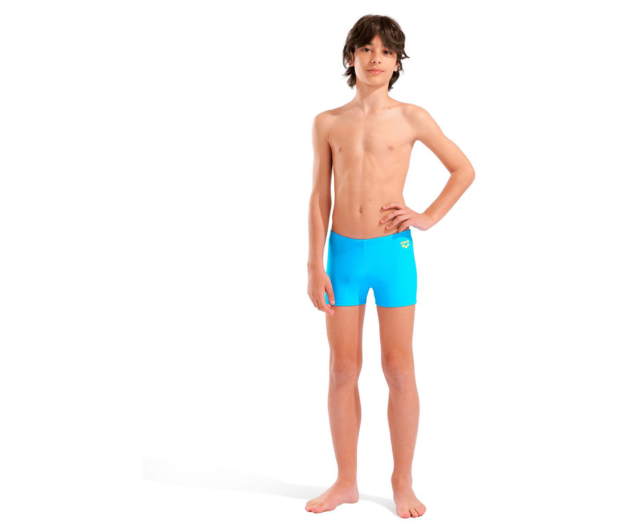 Młody mężczyzna stoi boso na białym tle, ubrany w niebieskie kąpielówki.