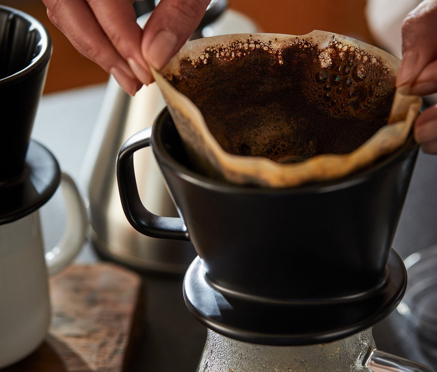 Kobieta umieszcza papierowy filtr w czarnym ręcznym filtrze do kawy, rozm. 2. Pod spodem znajduje się dzbanek do kawy, szklany.