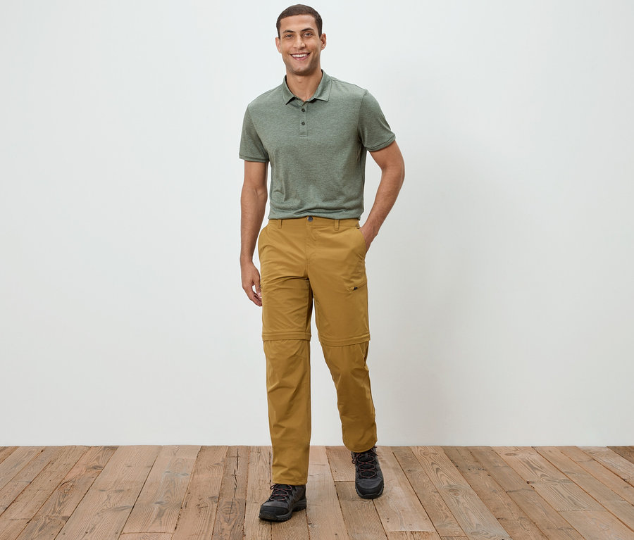 Mężczyzna stoi na tle białej ściany, ubrany w zieloną koszulkę polo i spodnie khaki.