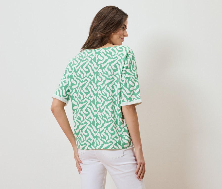 Widok z tyłu na kobietę z brązowymi włosami, ubraną w bluzkę z krótkim rękawem w zielono-biały wzór i białe dżinsy.