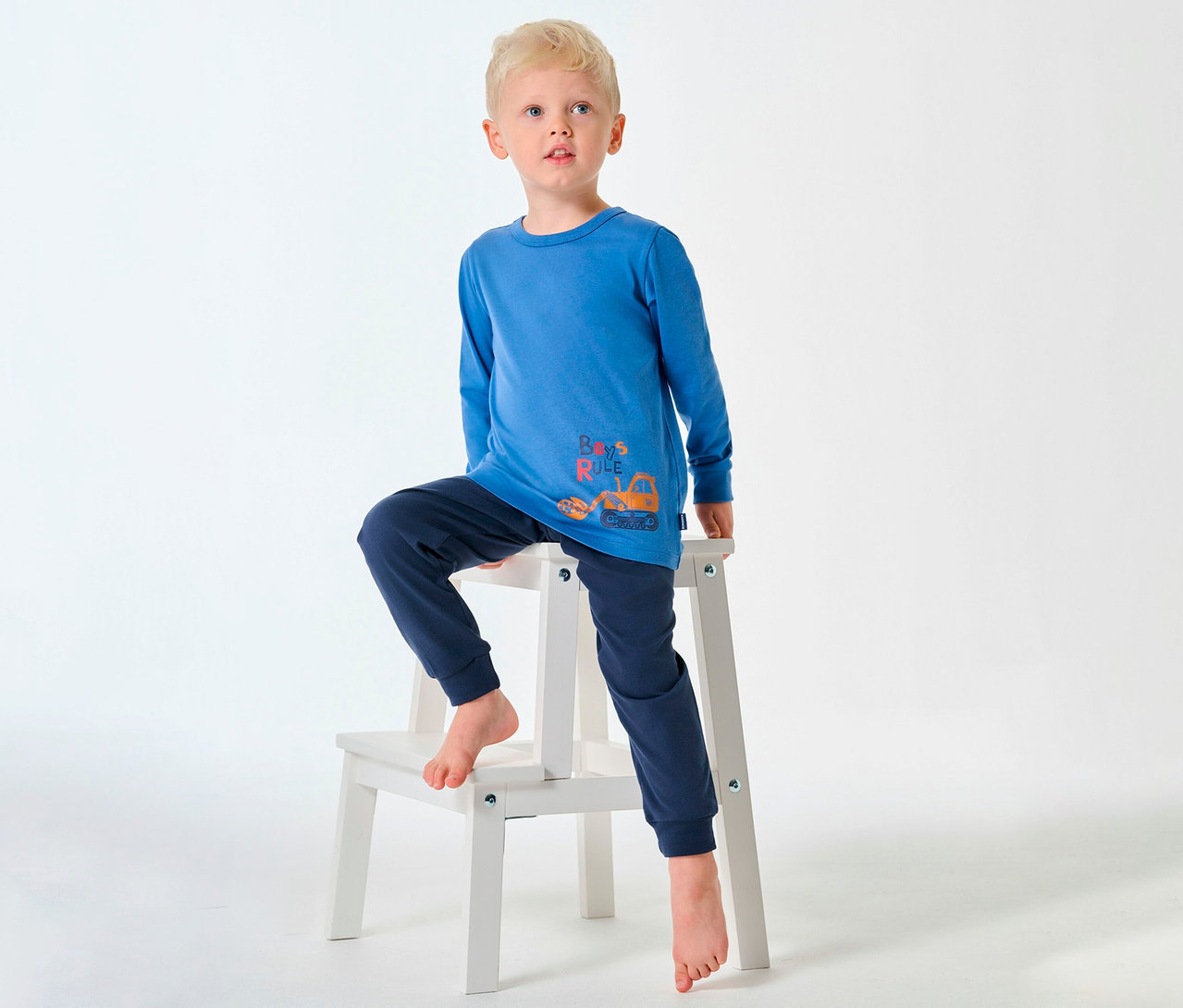Blondwłosy chłopiec w piżamie dziecięcej SCHIESSER siedzi boso na stołku.