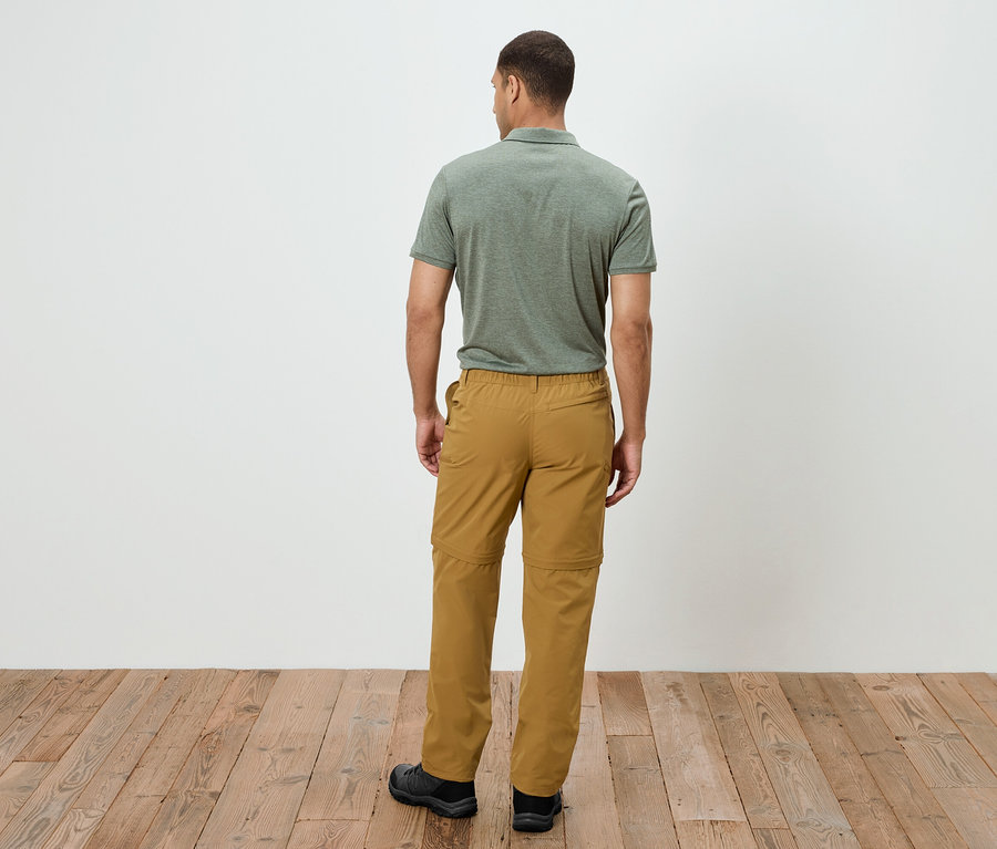 Mężczyzna stoi tyłem do kamery, ubrany w oliwkową koszulkę polo i złote spodnie outdoorowe z zamkiem błyskawicznym. Stoi na drewnianej podłodze.