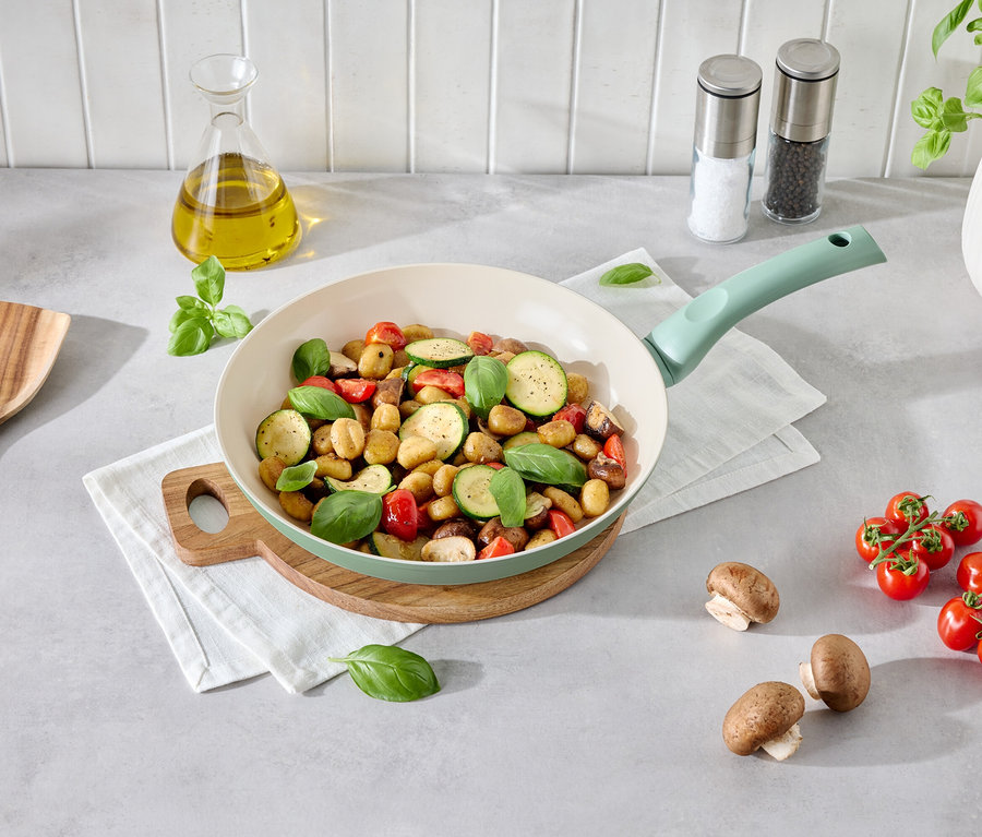 Gnocchi z warzywami na jasnozielonej patelni na drewnianym talerzu. Obok leżą pieczarki, pomidory, drewniana łyżka i butelka oleju.