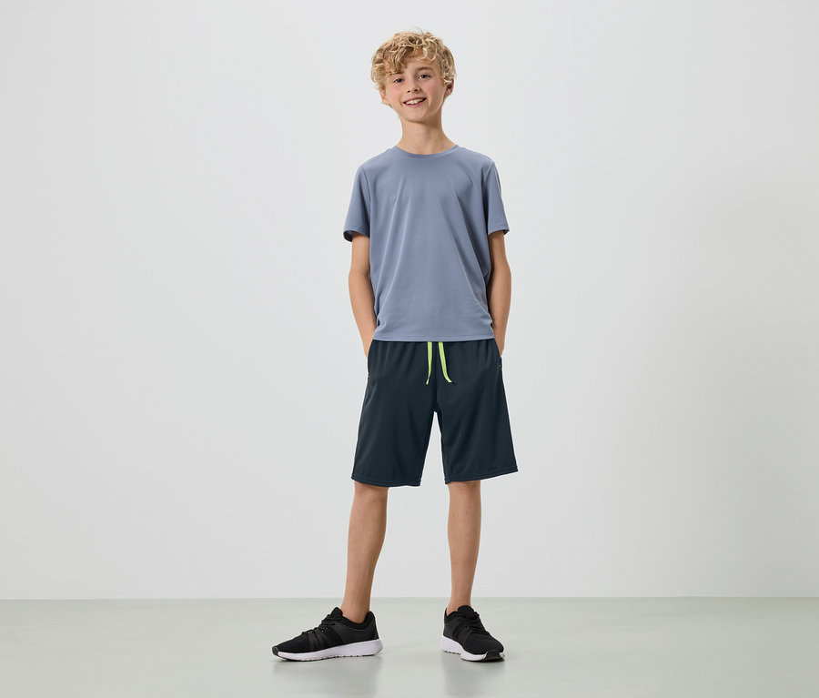 Młody mężczyzna z blond lokami stoi na białym tle w szarej koszulce, granatowych szortach i czarnych butach sportowych.