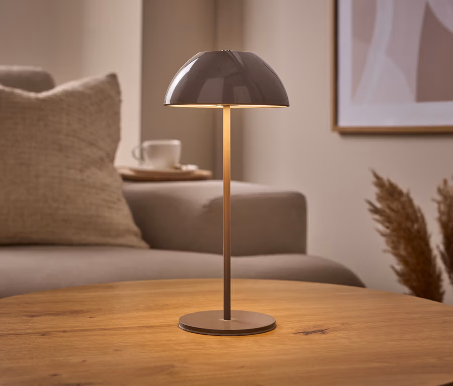 Akumulatorowa lampa stołowa LED, beżowa, z kopułowym kloszem stoi na drewnianym stole. W tle sofa, filiżanka i trawa pampasowa.