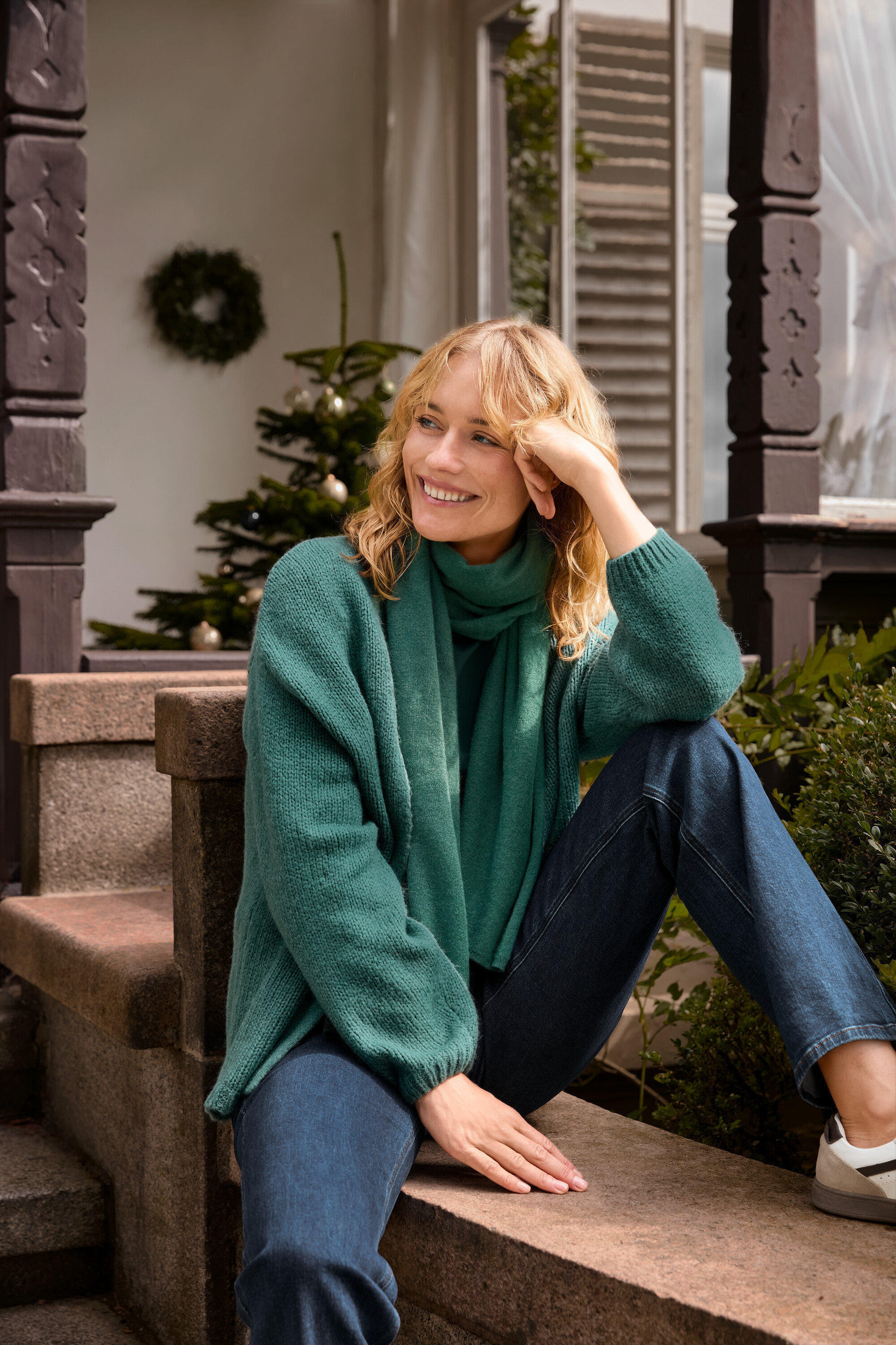 Blondwłosa kobieta siedzi na schodach, ma na sobie kardigan bez zapięcia o grubym splocie, zielony tkany szalik i spodnie dżinsowe typu bootcut – fason „Lou”.