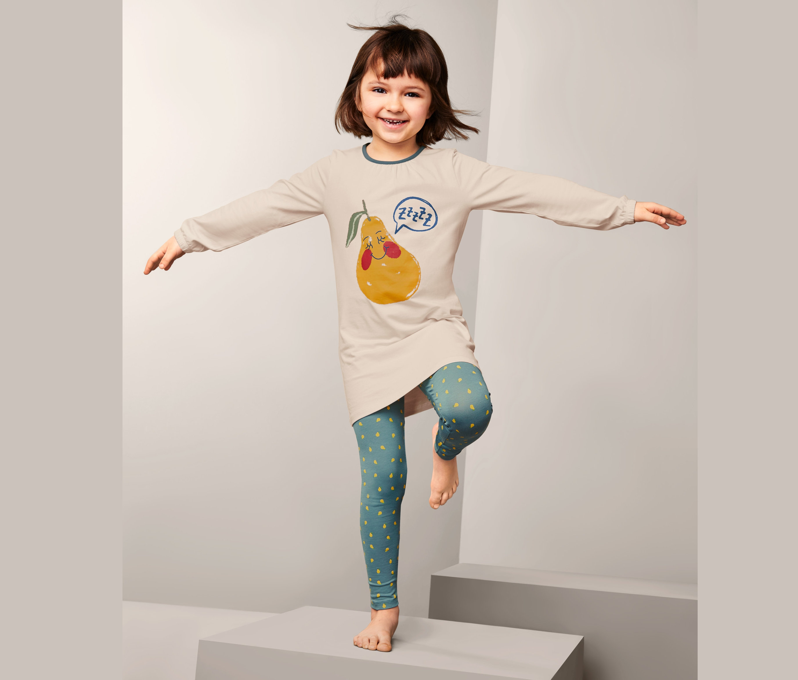 Dziewczynka stoi na jednej nodze w dziecięcej piżamie w gruszki.