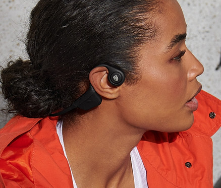 Kobieta siedzi obok roweru i trzyma kask, ma na uszach Słuchawki Bluetooth® z przewodnictwem kostnym.