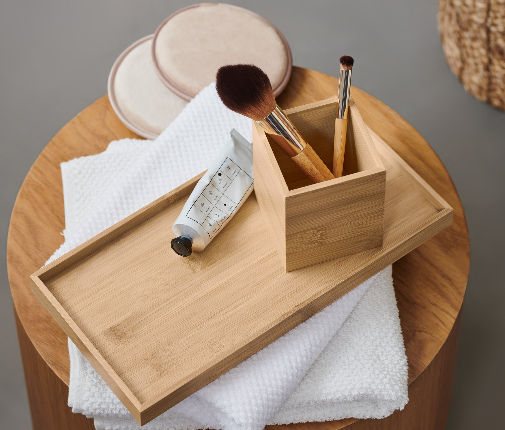 Na okrągłym stoliku leży dekoracyjna taca z pędzlami do makijażu, wacikami do demakijażu, ręcznikiem i tubką kremu.