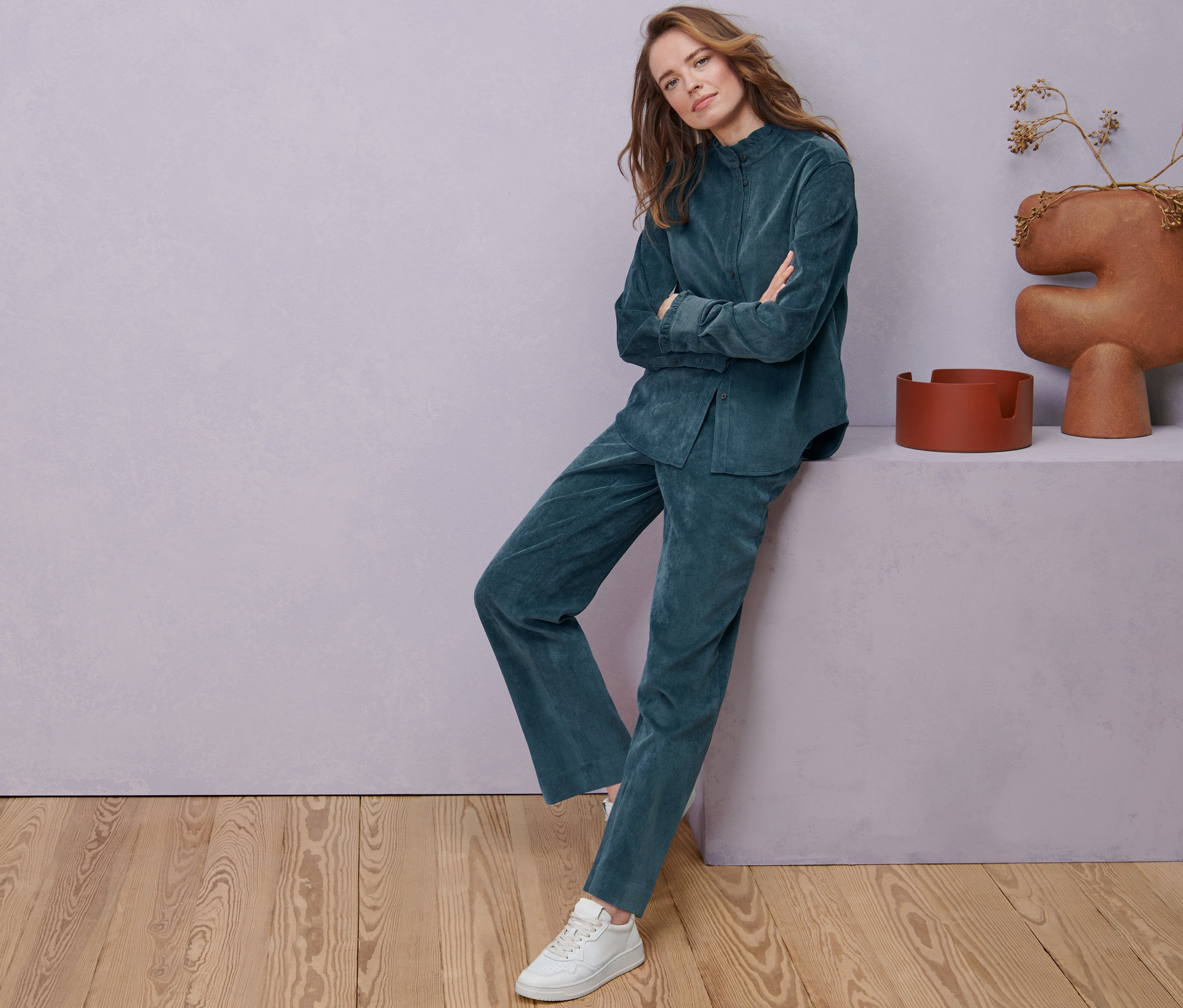 Kobieta pozuje w bluzce sztruksowej z falbanką i szaroniebieskich spodniach sztruksowych.