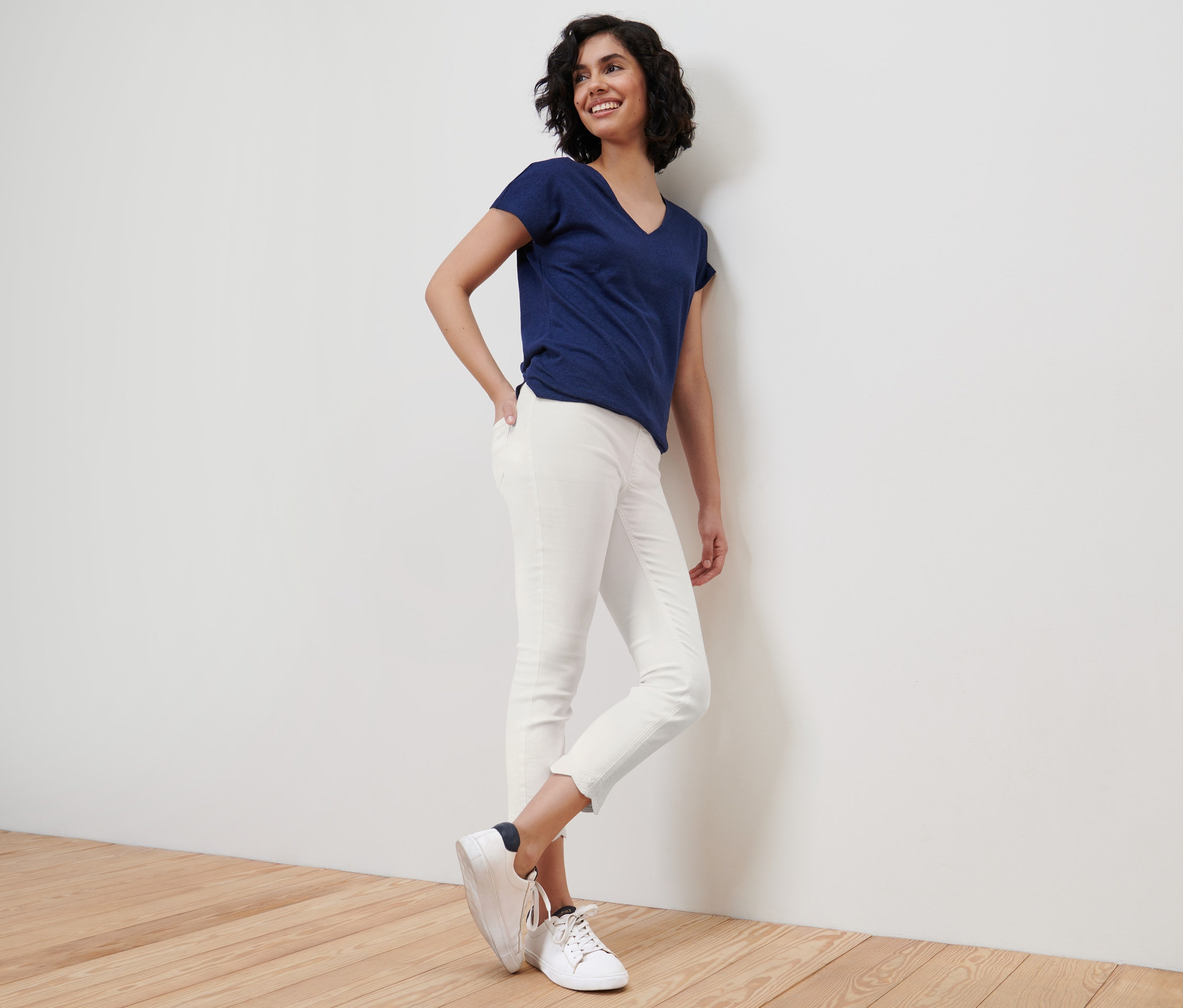 Uśmiechnięta kobieta opiera się o ścianę, ubrana w niebieski T-shirt i białe tregginsy o długości 7/8 z ażurowym haftem.