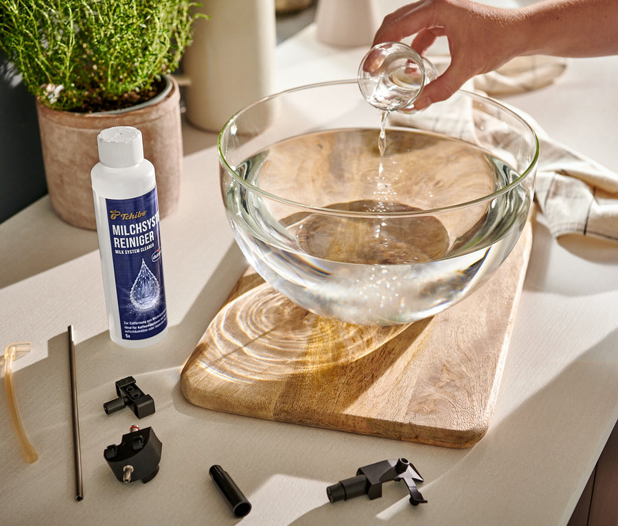 Ręka nalewa klarowny płyn z miarki do szklanej miski z wodą. Obok stoi środek do czyszczenia układu mlecznego durgol® i części ekspresu do kawy.