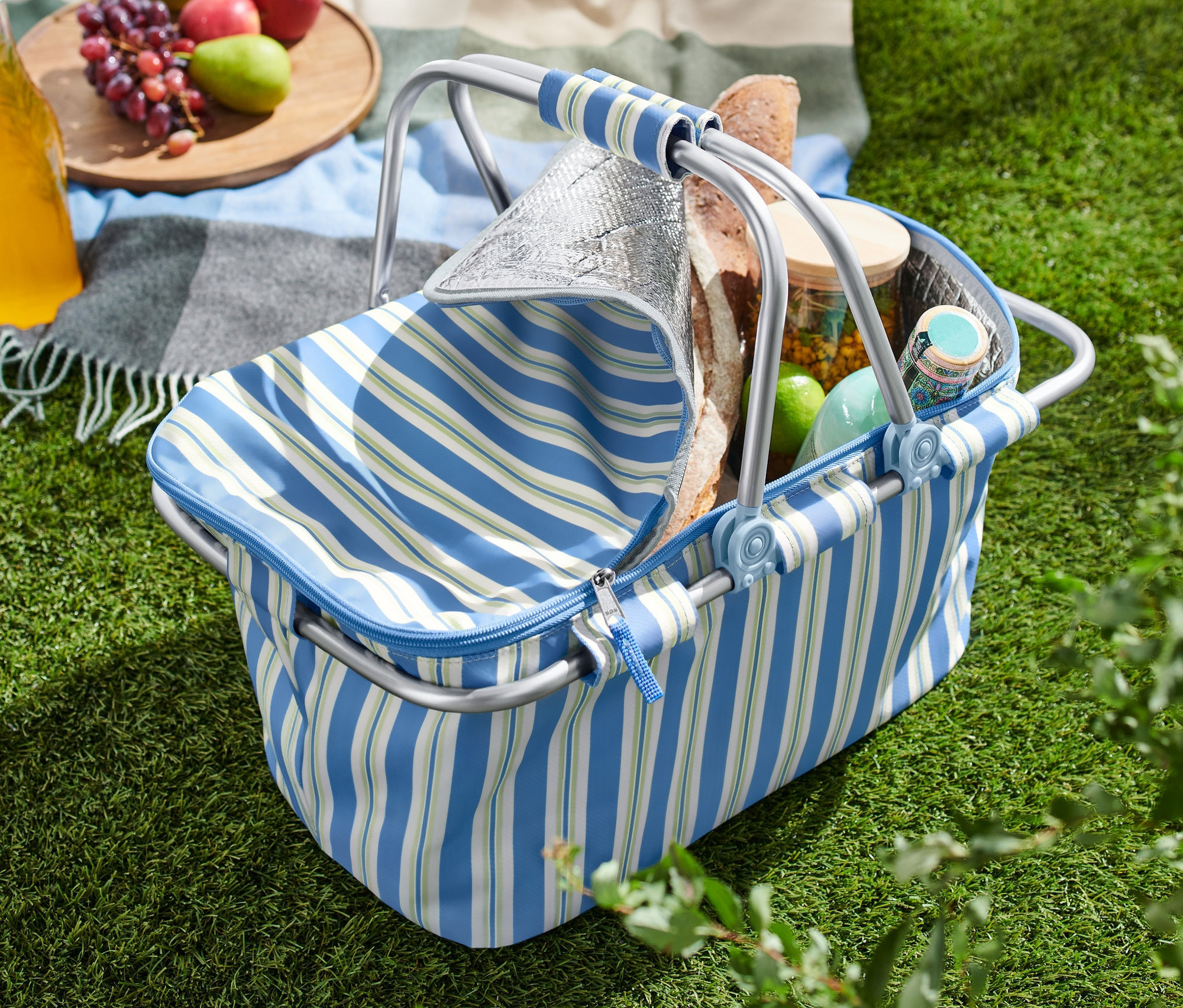 Otwarty koszyk termiczny na trawie obok koca z jabłkami, winogronami i napojem.