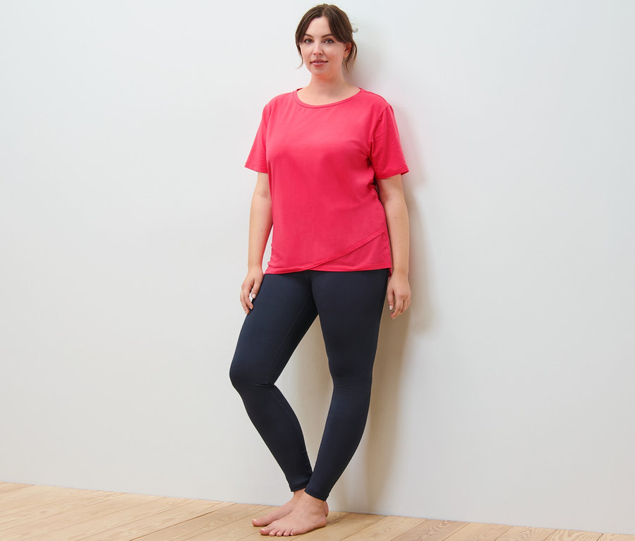 Kobieta stoi oparta o ścianę, ubrana w koszulkę sportową i legginsy sportowe, granatowe.