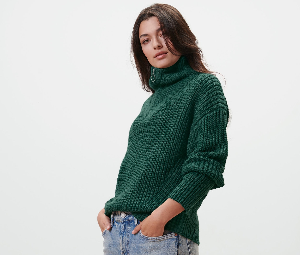 Kobieta pozuje w zielonym grubym golfie z rozpinanym kołnierzem i jeansach.