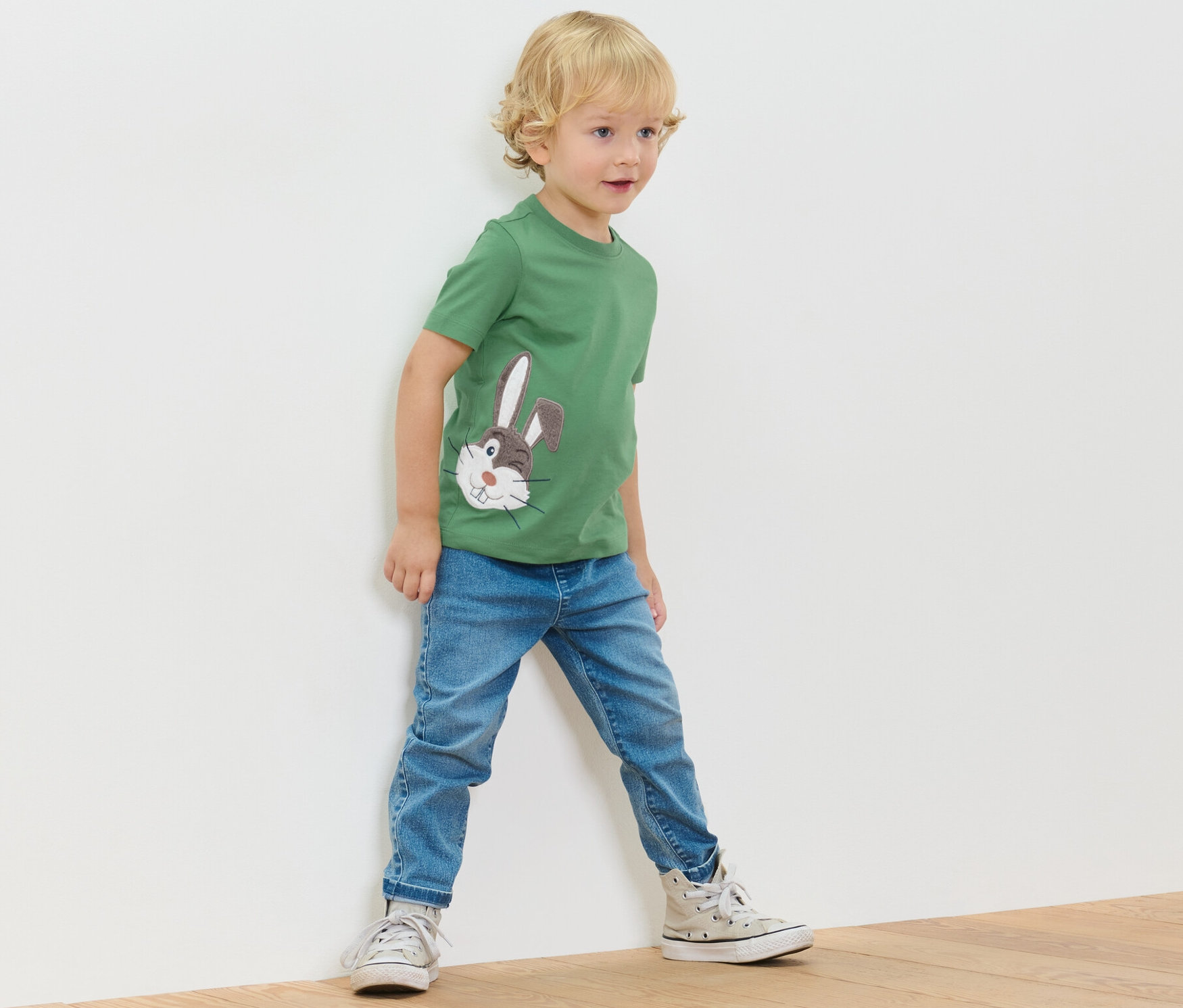 Chłopiec stoi przy ścianie, ubrany w zieloną koszulkę z aplikacją i dżinsy.