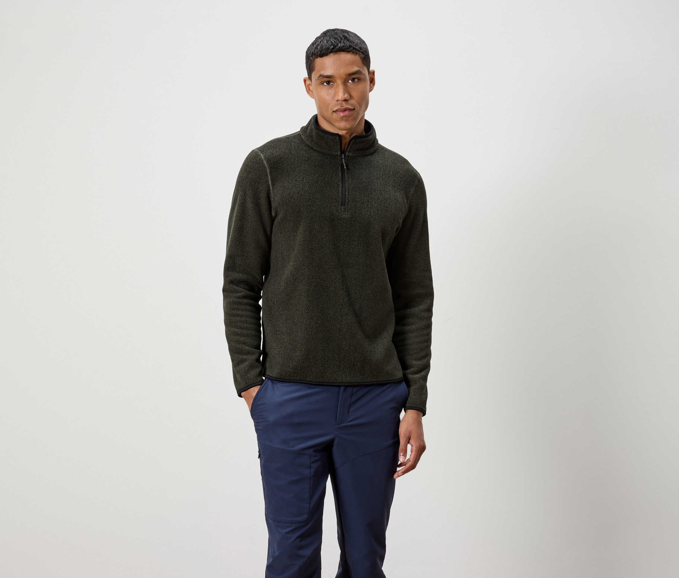 Mężczyzna stoi, ubrany w oliwkowy sweter na zamek i niebieskie spodnie.