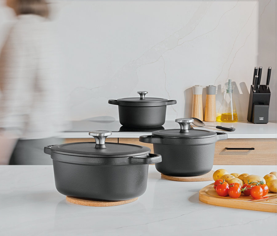Trzy czarne garnki z pokrywkami na blacie kuchennym, obok deska do krojenia z pomidorami i ziemniakami.