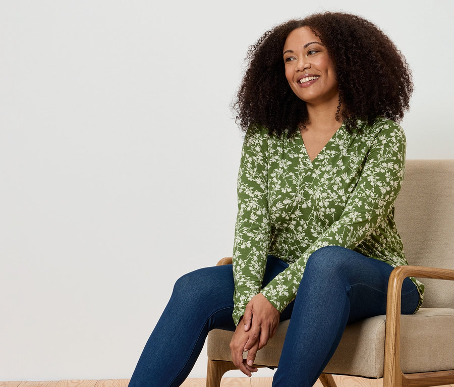Kobieta siedzi w fotelu. Ma na sobie zieloną bluzkę w kwiaty i dżinsy.
