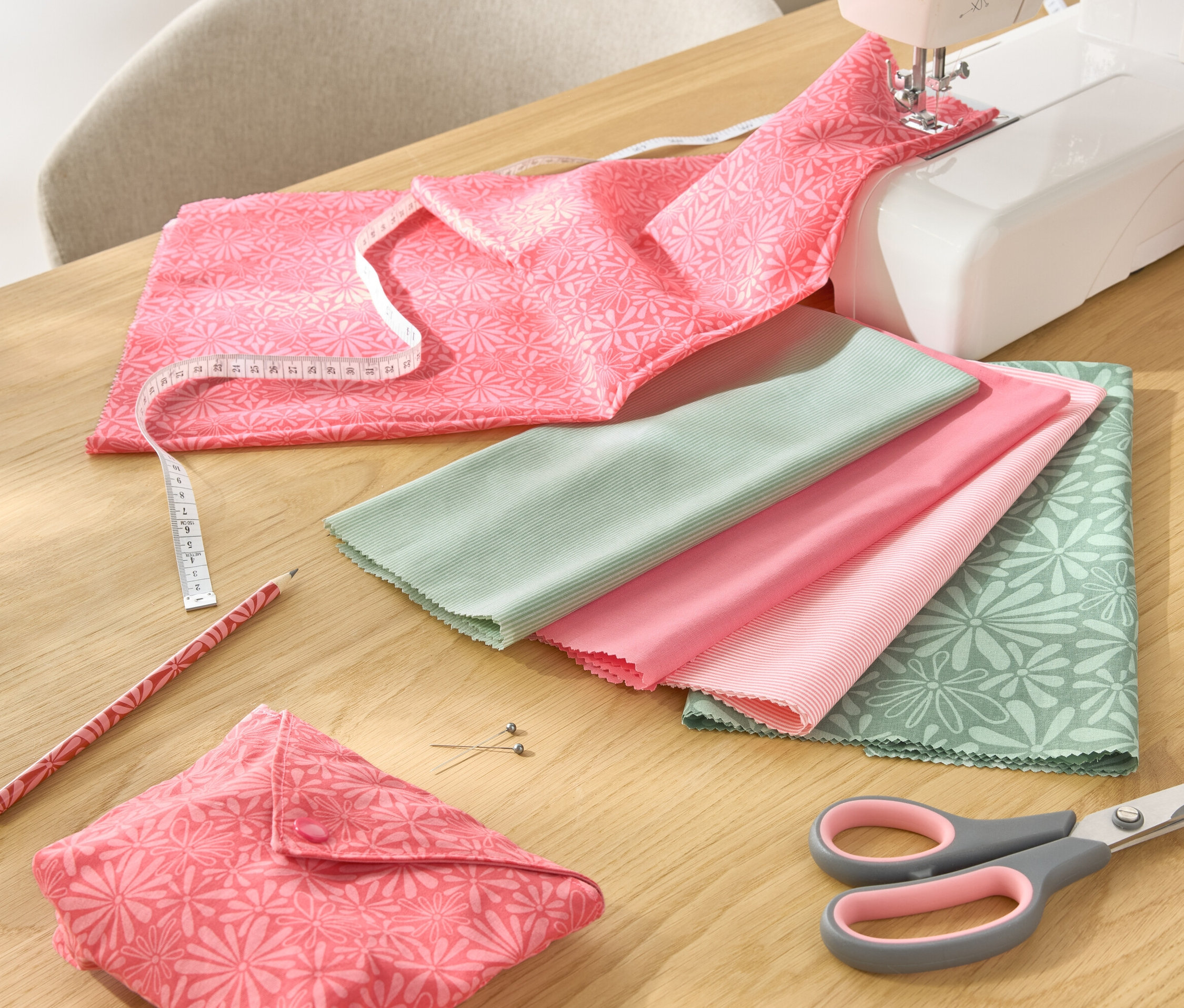 Na stole leży 5-częściowy zestaw materiałów, maszyna do szycia, nożyczki, ołówek i miarka krawiecka.