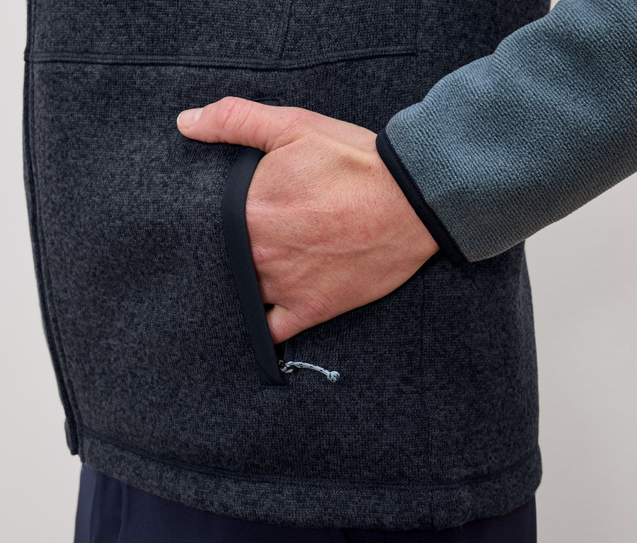 Mężczyzna wkłada rękę do kieszeni szaro-niebieskiej kurtki polarowej.