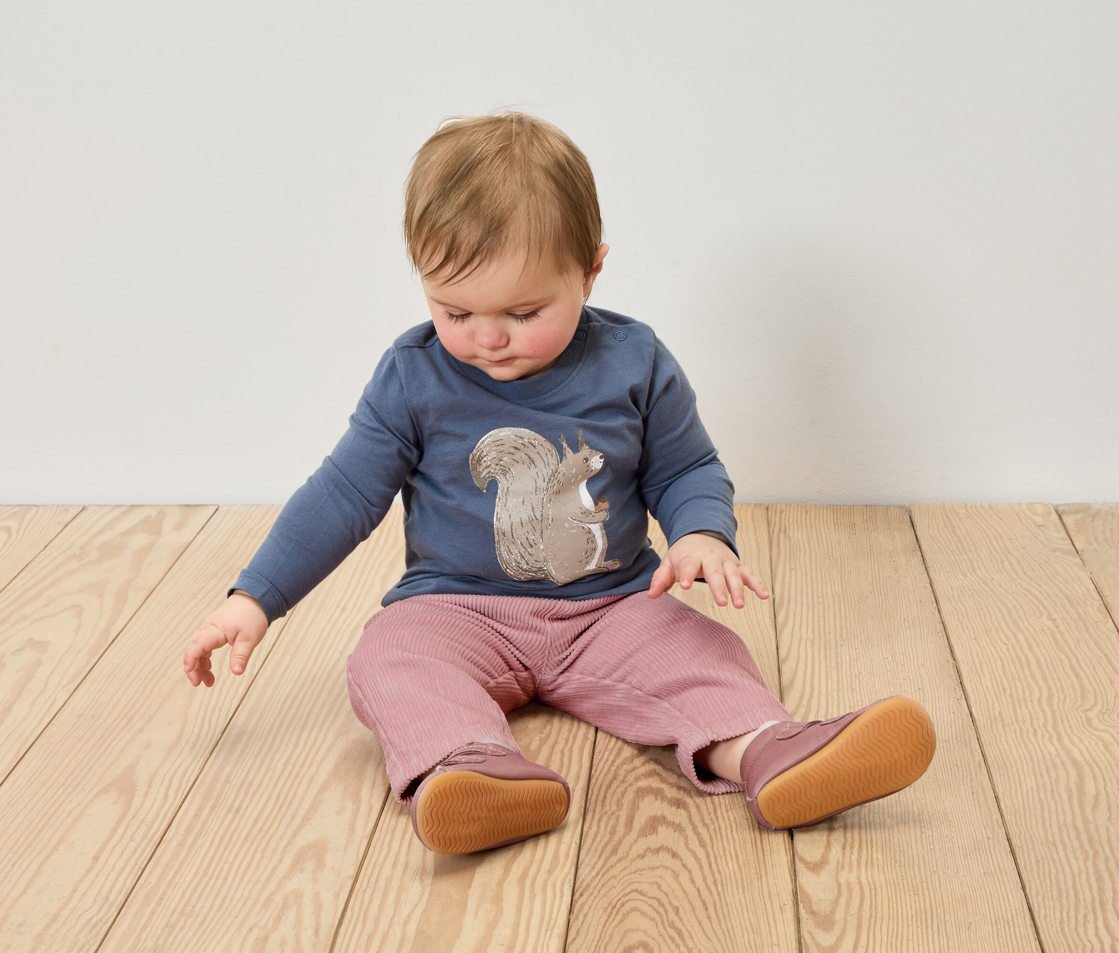 Niemowlę siedzi na drewnianej podłodze, ubrane w niebieską koszulkę z nadrukiem wiewiórki, różowe spodnie i skórzane buciki.