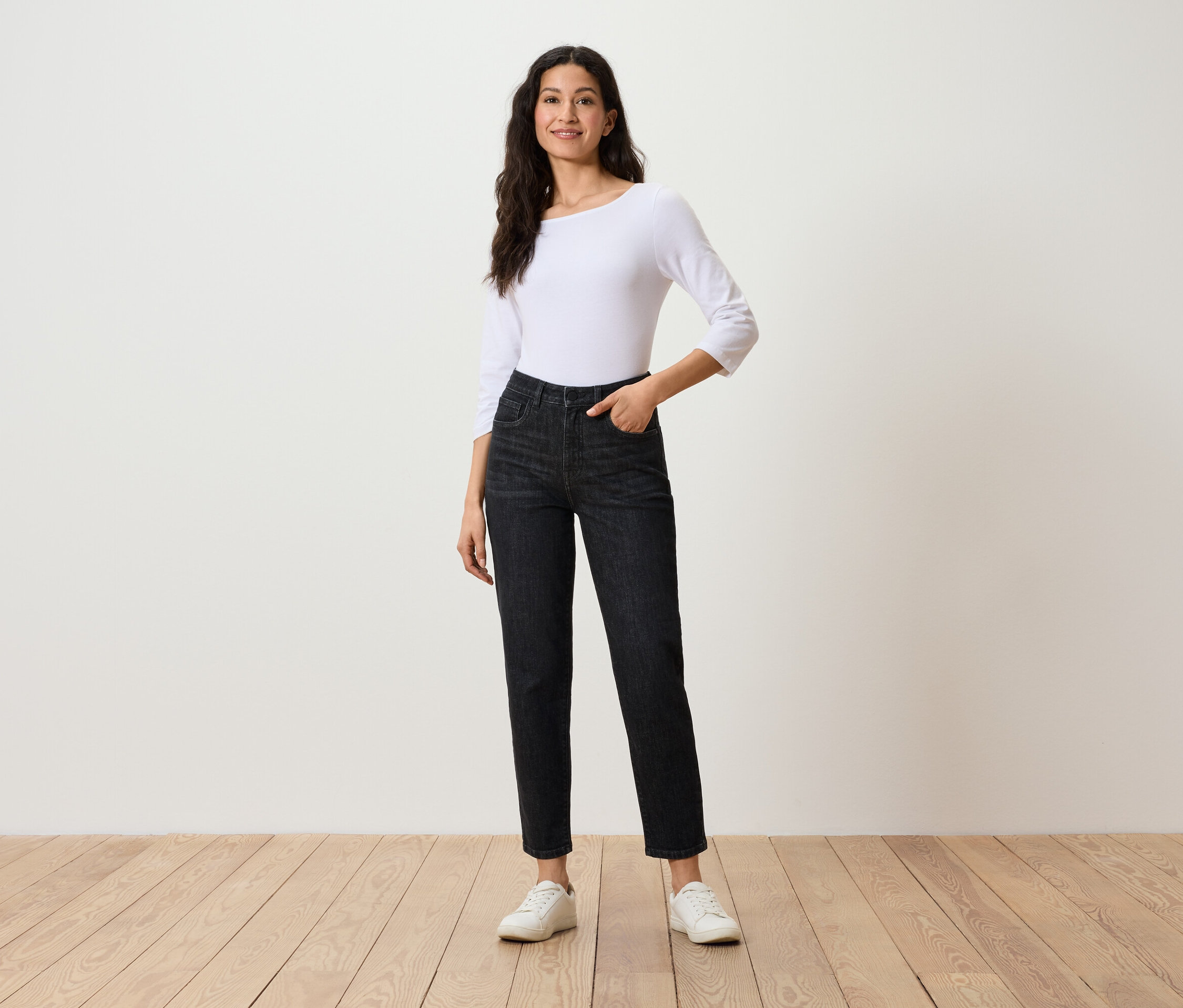 Kobieta pozuje w czarnych dżinsach, białym topie z rękawami trzy czwarte i białych tenisówkach.