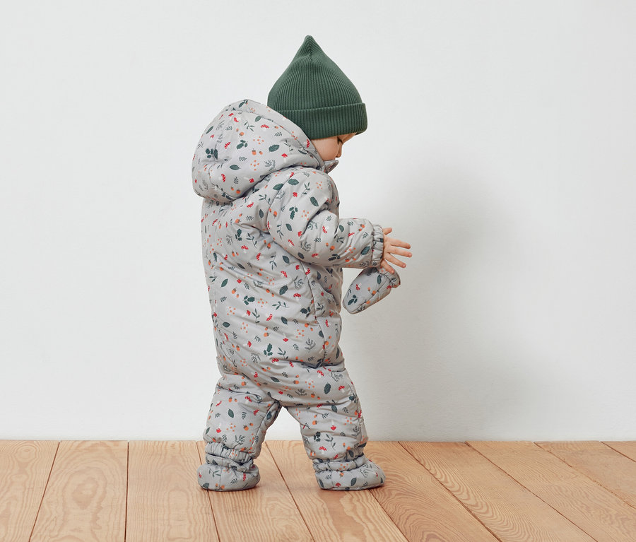 Małe dziecko w szarym kombinezonie w kwiatowy wzór i zielonej czapce.