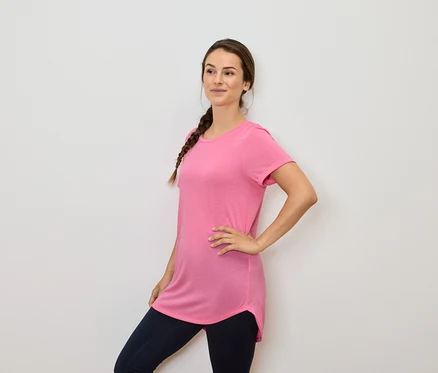 Kobieta z brązowymi włosami w warkoczu pozuje w różowym t-shircie i granatowych legginsach.