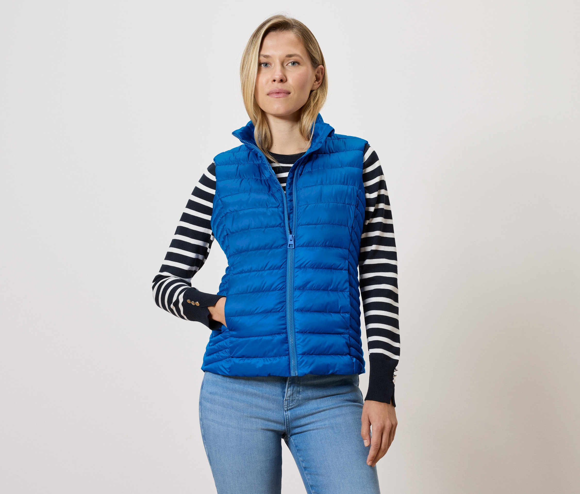 Kobieta w niebieskiej pikowanej kamizelce na zamek błyskawiczny, swetrze w czarno-białe paski i niebieskich dżinsach.