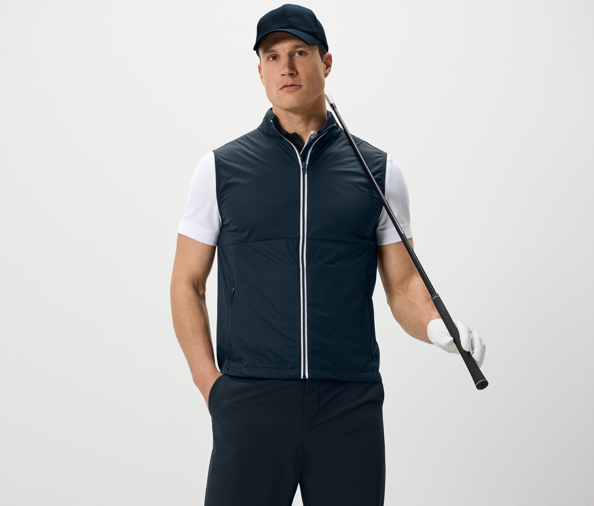 Mężczyzna ma na sobie kamizelkę softshellową i czapkę z daszkiem, trzyma kij golfowy i stoi na białym tle.