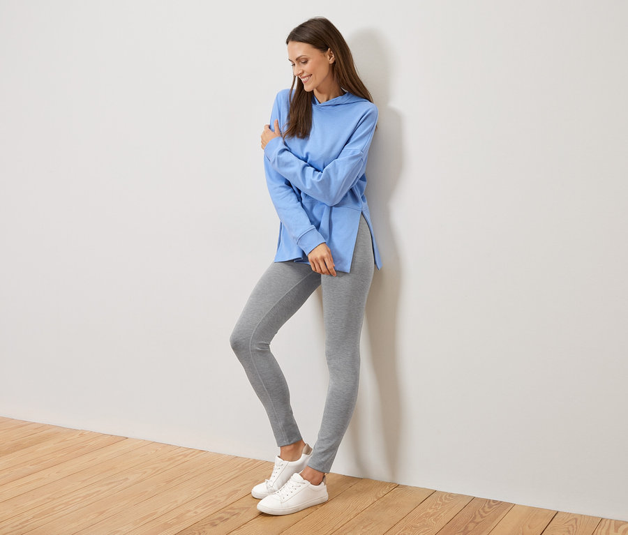 Kobieta opiera się o ścianę, ma na sobie jasnoniebieską bluzę z kapturem z rozcięciami po bokach, szare legginsy i białe trampki.
