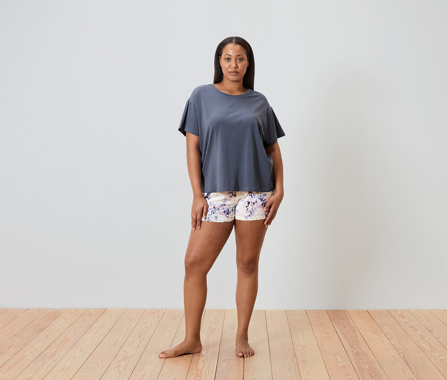 Kobieta stoi boso na drewnianej podłodze, ubrana w szarą koszulkę i szorty w kwiaty.