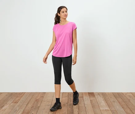 Kobieta stoi na drewnianej podłodze, ubrana w różowy T-shirt, czarne legginsy Capri i czarne tenisówki.