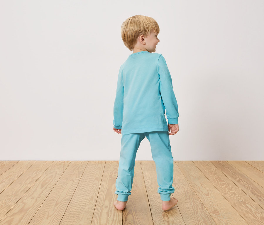 Chłopiec od tyłu, ubrany w jasnoniebieską piżamę, stoi na drewnianej podłodze przed białą ścianą.