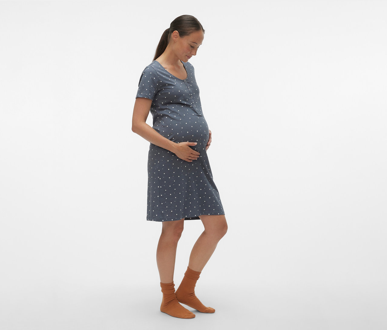 Kobieta w ciąży z profilu ma na sobie niebieską koszulę nocną MAMALICIOUS „MLMIRA” w białe kropki i brązowe skarpetki.