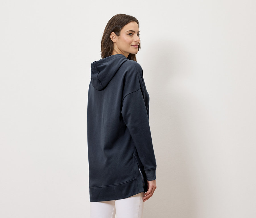 Kobieta ubrana w długą granatową bluzę z kapturem i białe spodnie, widziana z boku.