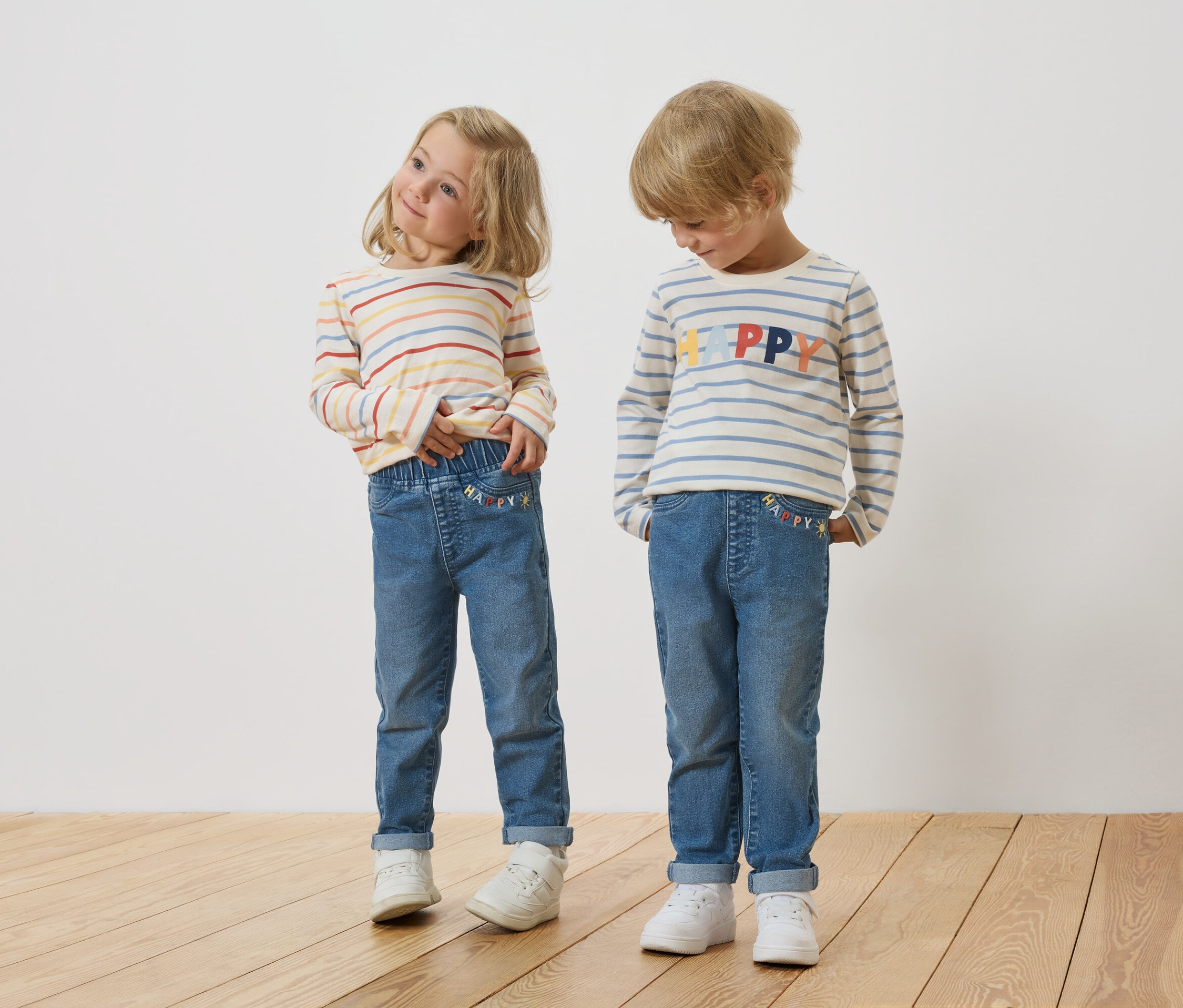 Dwoje blondwłosych dzieci, dziewczynka i chłopiec, stoi obok siebie, ubrani w bluzki z długim rękawem w paski i dżinsy.