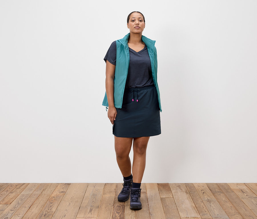 Kobieta stoi na tle białej ściany, ubrana w granatową spódnicę, granatowy T-shirt, turkusową kamizelkę i granatowe buty trekkingowe.