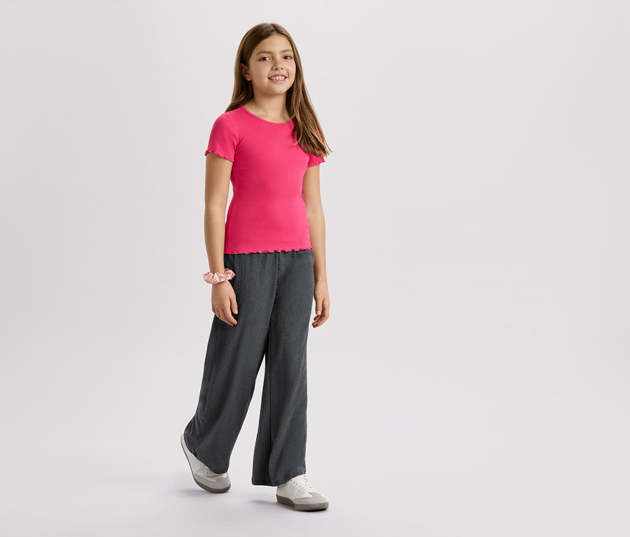 Dziewczyna w różowym T-shircie, szarych spodniach i trampkach na białym tle.