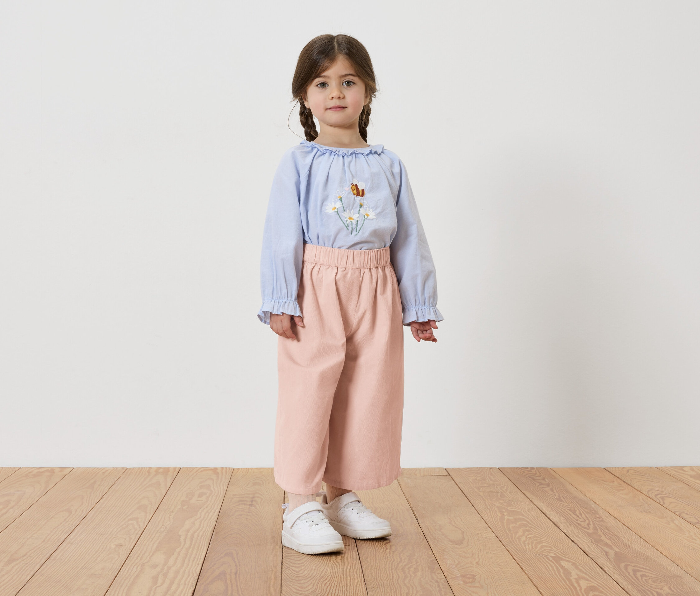 Dziewczynka z warkoczami ma na sobie jasnoniebieską bluzkę z haftem kwiatowym, różowe spodnie i białe tenisówki.