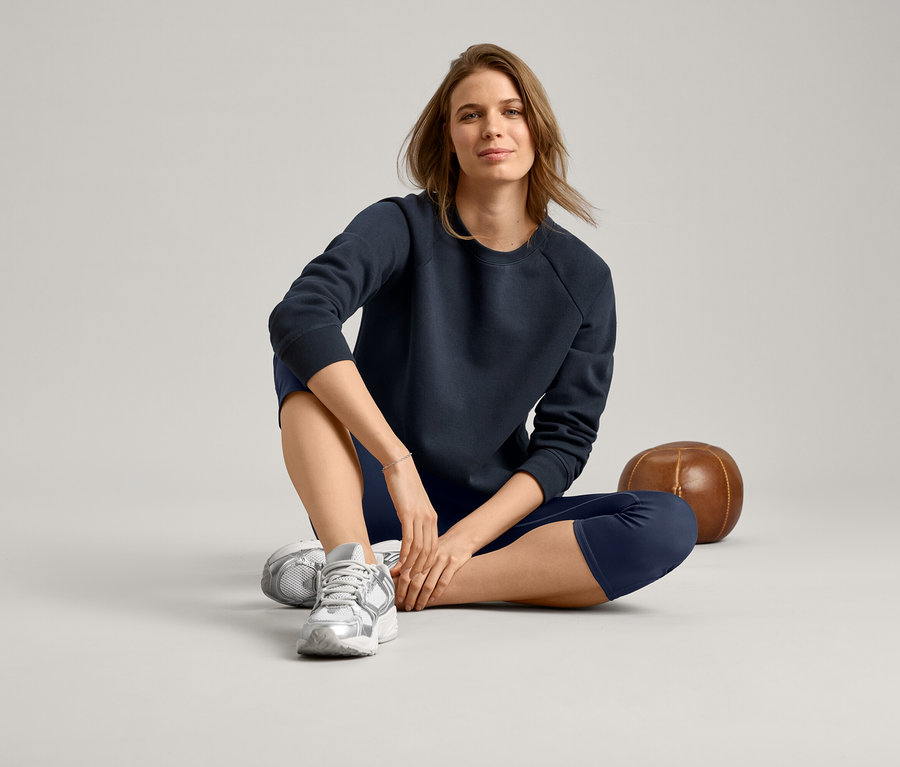 Kobieta siedzi po turecku, ma na sobie granatową bluzę, granatowe legginsy i srebrne buty sportowe.