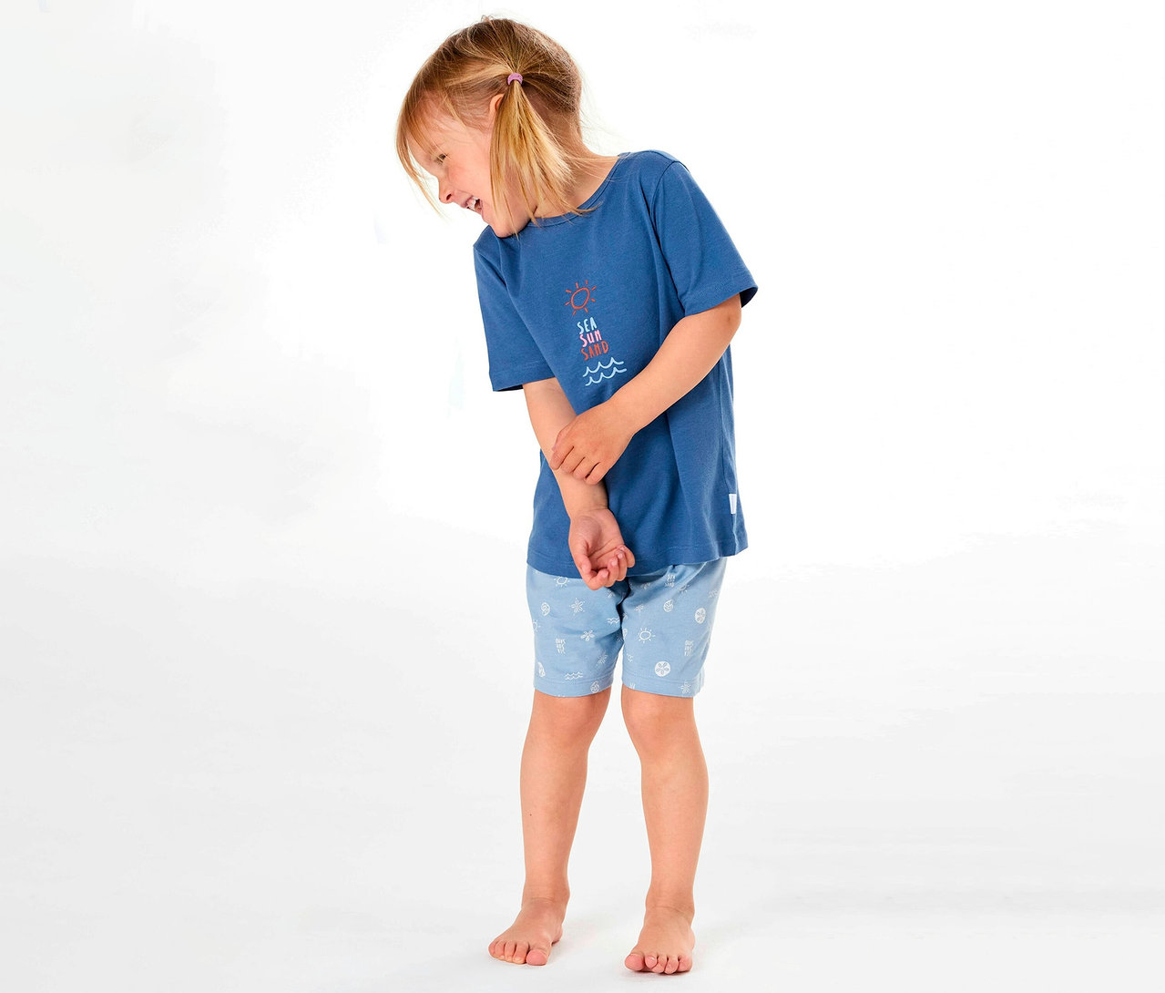 Dziewczynka w krótkiej piżamie dziecięcej SCHIESSER z niebieską koszulką i wzorzystymi szortami, uśmiecha się w bok.