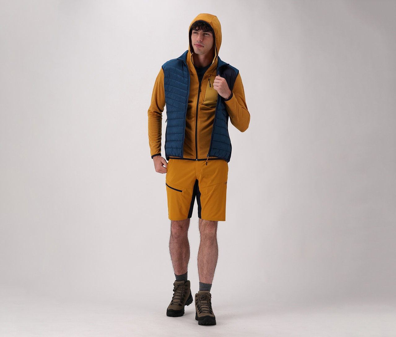 Mężczyzna ma na sobie bluzę CMP z kapturem, kamizelkę hybrydową CMP, szorty i buty na jasnym tle.