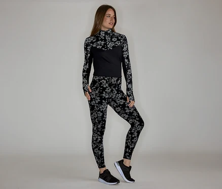 Kobieta w kwiecistej bluzce z długim rękawem i legginsach, do tego czarne buty sportowe.