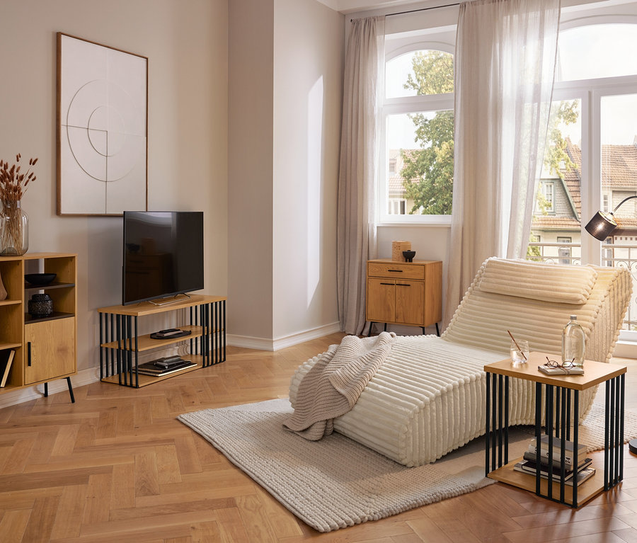 Nowoczesny salon z kremowym szezlongiem na jasnym dywanie, drewnianą szafką, szafką RTV i stolikiem bocznym. Duże okna z zasłonami wychodzą na zewnątrz.