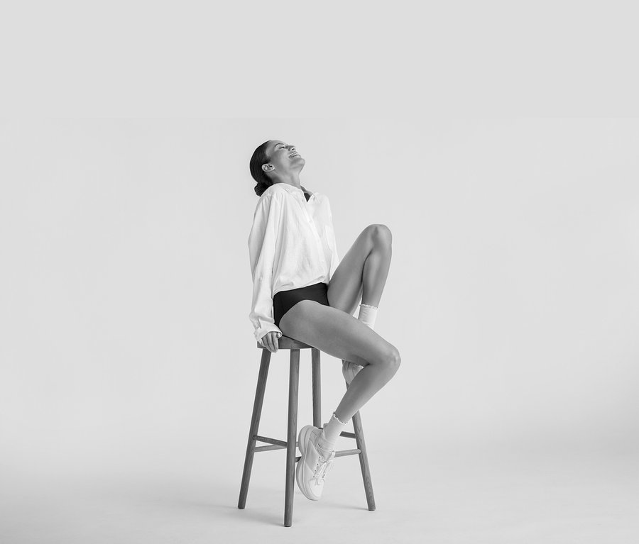 Kobieta siedzi uśmiechnięta na stołku, ma na sobie białą Długą lnianą bluzkę, czarną bieliznę, białe skarpetki i białe trampki.