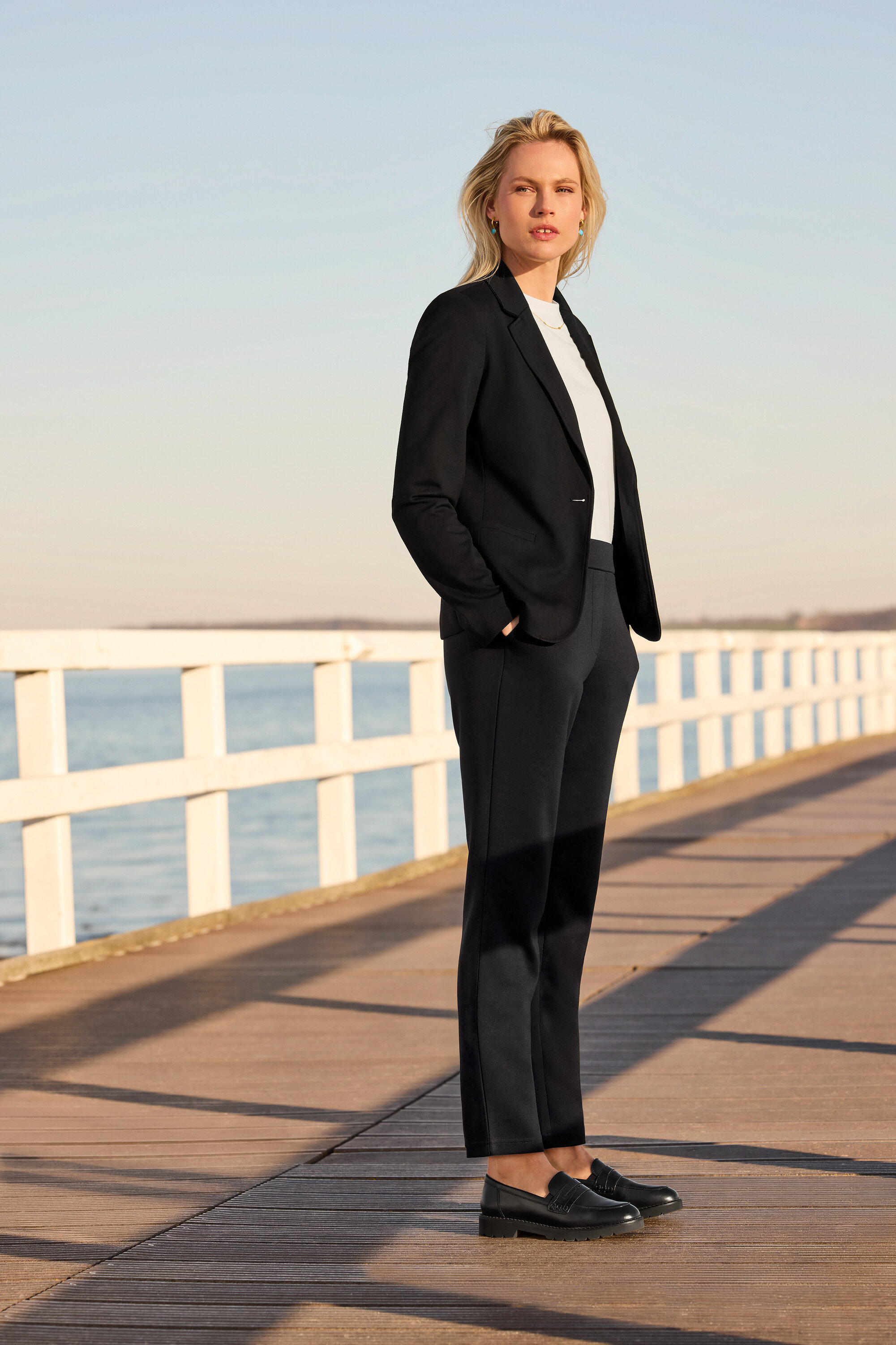 Kobieta stojąca na molo ma na sobie spodnie dresowe, białą koszulkę, żakiet z dżerseju i skórzane loafersy. Nosi kolczyki kreole z kamiennym wisiorkiem i naszyjnik z kryształkami Navette.